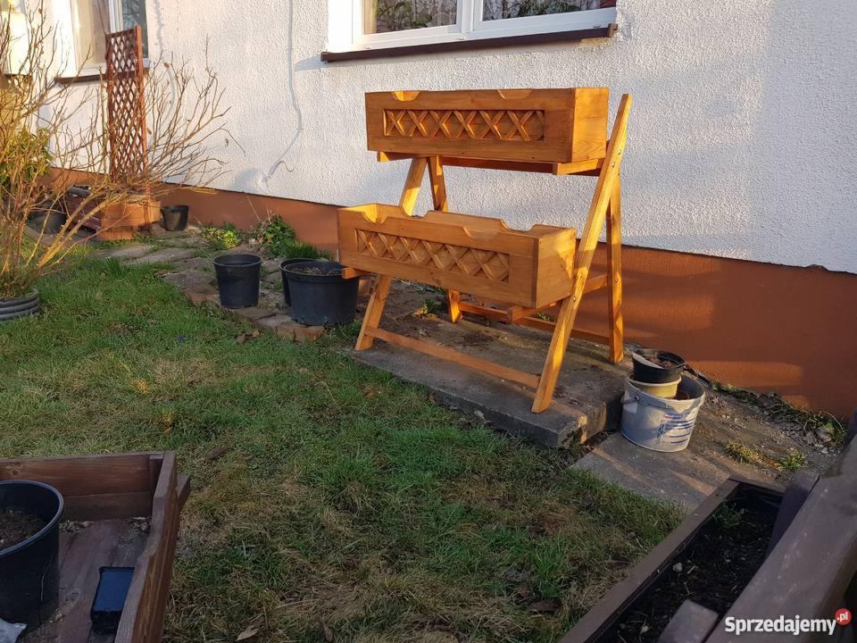 Dwupoziomowy, drewniany kwietnik z ozdobnym ażurem w kształcie krzyżyków, ustawiony na trawniku przed białym domem z brązową podmurówką, obok doniczki i wiadro z ziemią.