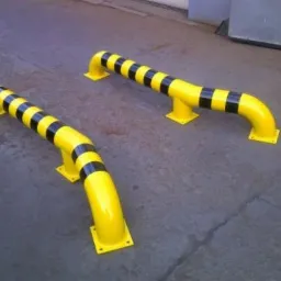 Dwie żółte, stalowe bariery ochronne z czarnymi pasami, zamontowane na szarym betonowym podłożu w hali.