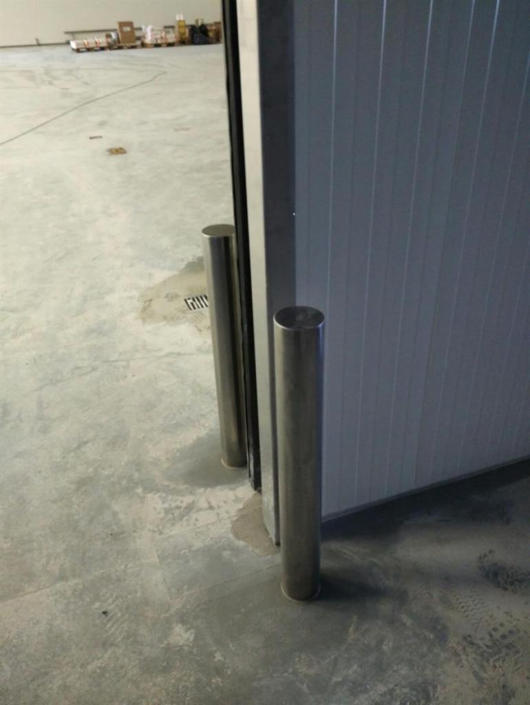 Dwie pionowe, cylindryczne poręcze ze stali nierdzewnej przy szarej ścianie z pionowymi panelami w surowym wnętrzu hali.