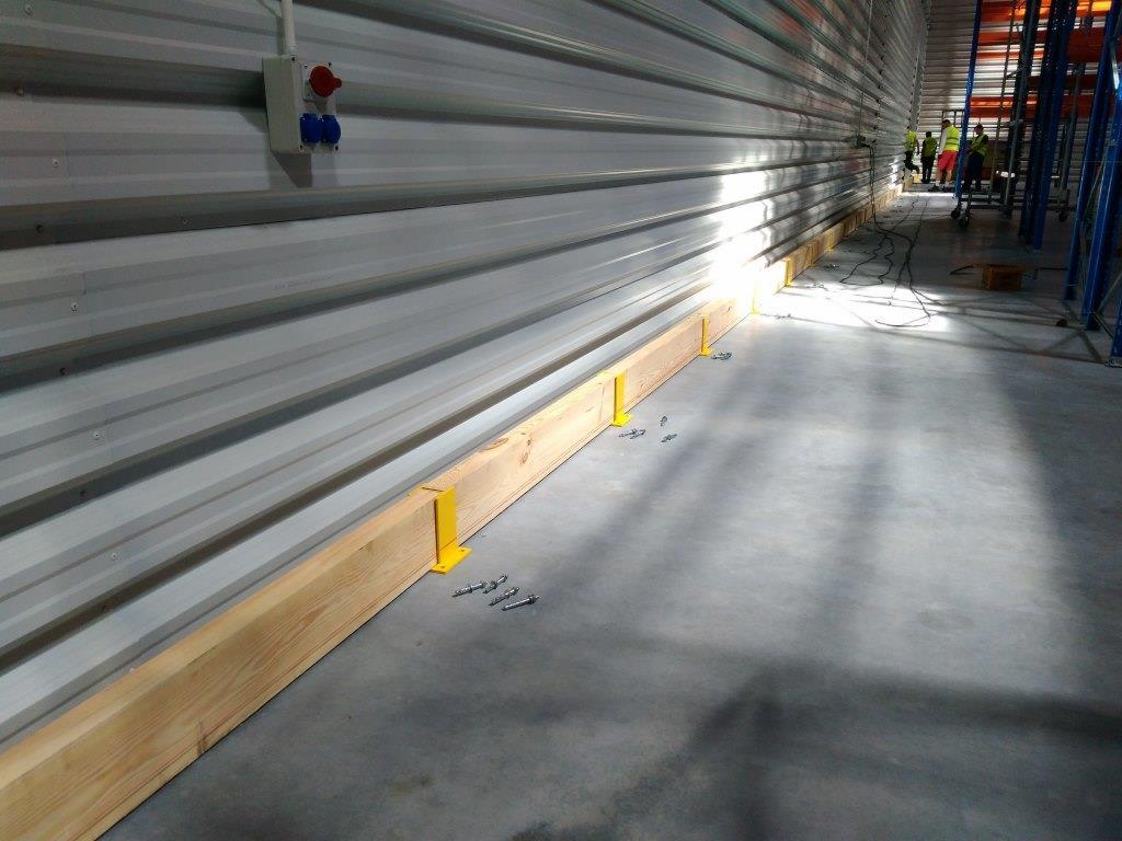 Montaż drewnianej listwy przypodłogowej w hali magazynowej, z widocznymi żółtymi wspornikami i śrubami na betonowej posadzce, przy metalowej ścianie z gniazdem elektrycznym.