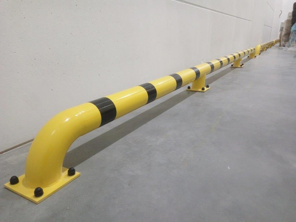 Długa, żółta poręcz zabezpieczająca z czarnymi pasami, zamocowana do podłogi w hali przemysłowej, widok z perspektywy podłogi.