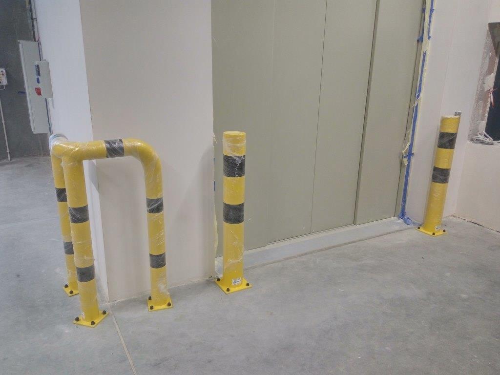 Żółte odbojniki rurowe z czarnymi pasami, zabezpieczone folią ochronną, zamontowane przy ścianach i drzwiach w surowym wnętrzu.