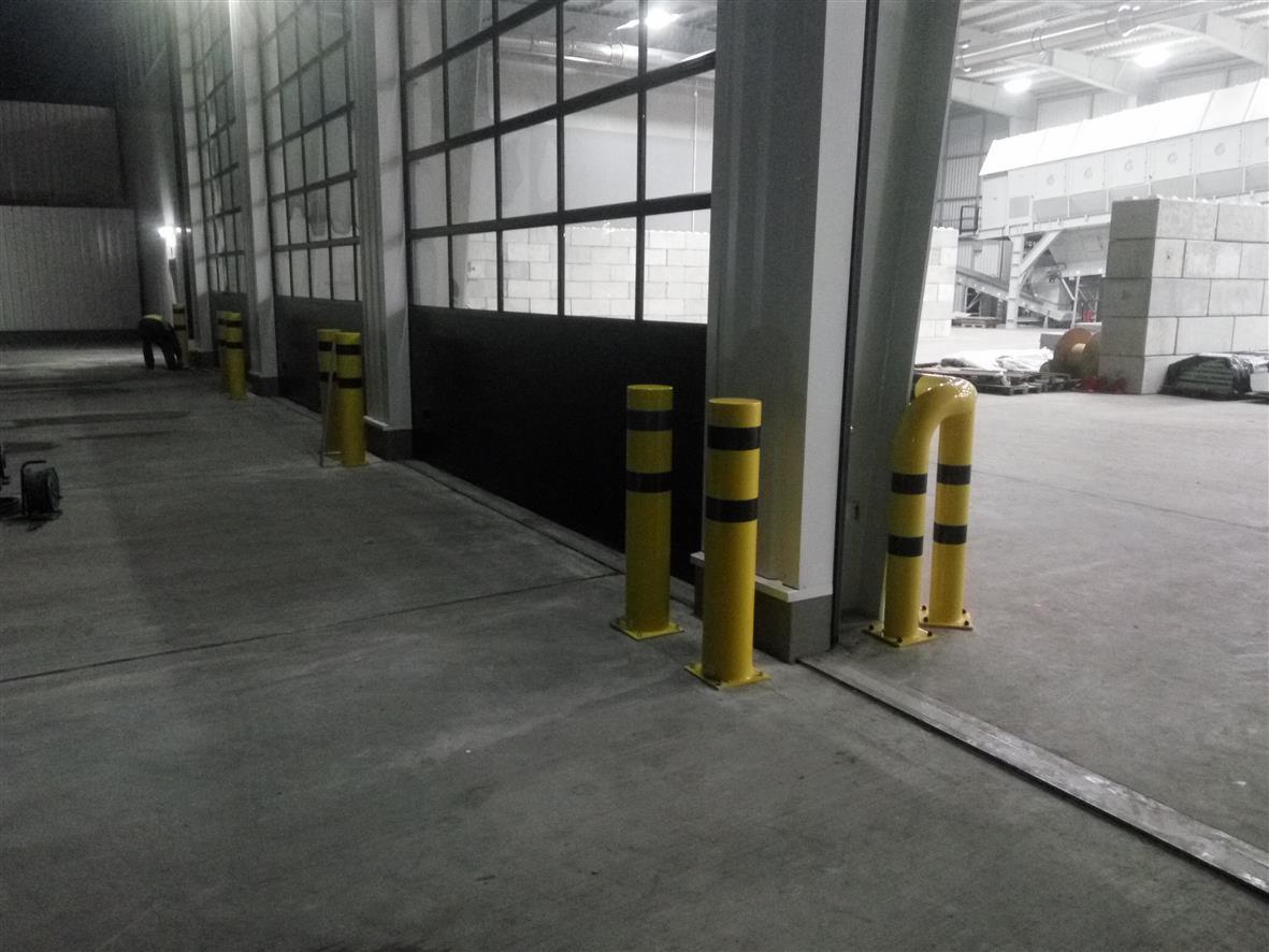 Żółte słupki ochronne z czarnymi pasami, ustawione wzdłuż dużej, przeszklonej, przesuwnej bramy przemysłowej w hali magazynowej.