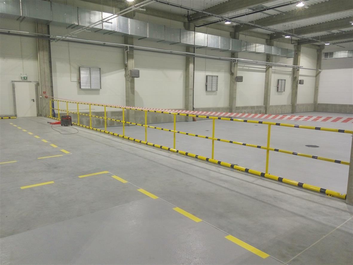 Żółto-czarne poręcze ochronne w hali magazynowej, widoczne oznaczenia na podłodze i sprzęt spawalniczy w tle.