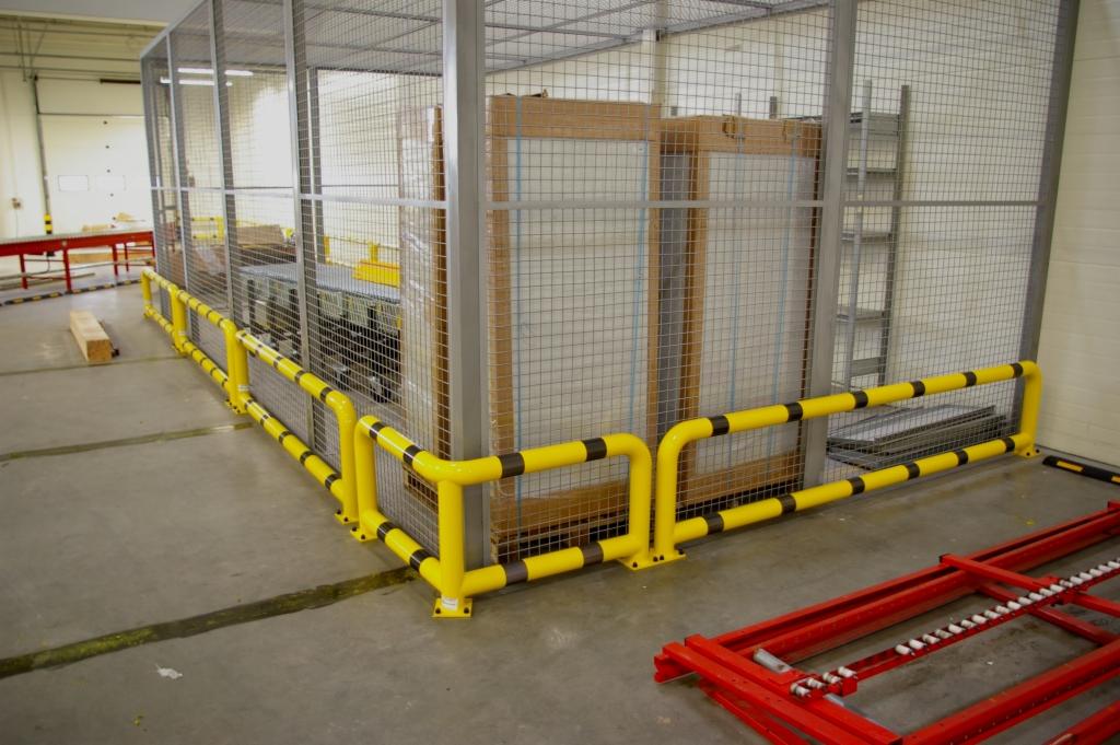 Klatka ochronna z siatki, zabezpieczona żółto-czarną poręczą rurową, w hali magazynowej.