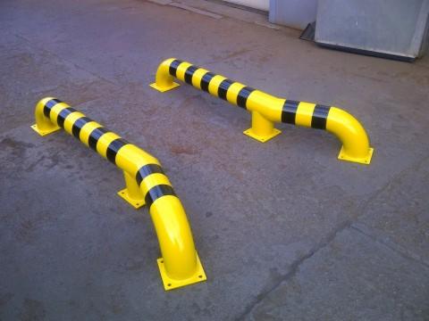 Dwie żółte, stalowe bariery ochronne z czarnymi pasami, zamontowane na szarym betonowym podłożu w hali.