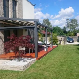 Nowoczesna altana w kolorze antracytowym z drewnianym tarasem, czerwonym klonem palmowym w donicy i zadbanym trawnikiem.