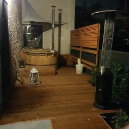 EWA WARDAK - Drewniany taras wieczorową porą z beczką kąpielową, dekoracyjnym lampionem i promiennikiem ciepła, oświetlony delikatnym światłem.