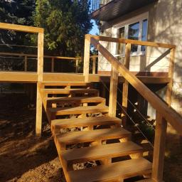 EWA WARDAK - Drewniane schody prowadzące na podwyższony taras z balustradą z cienkich linek, widok z dołu na tle budynku z balkonem i drzew