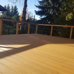 EWA WARDAK - Jasna, nowa powierzchnia tarasu z desek kompozytowych z drewnianą balustradą i stalowymi linkami, w tle drzewa i błękitne niebo.