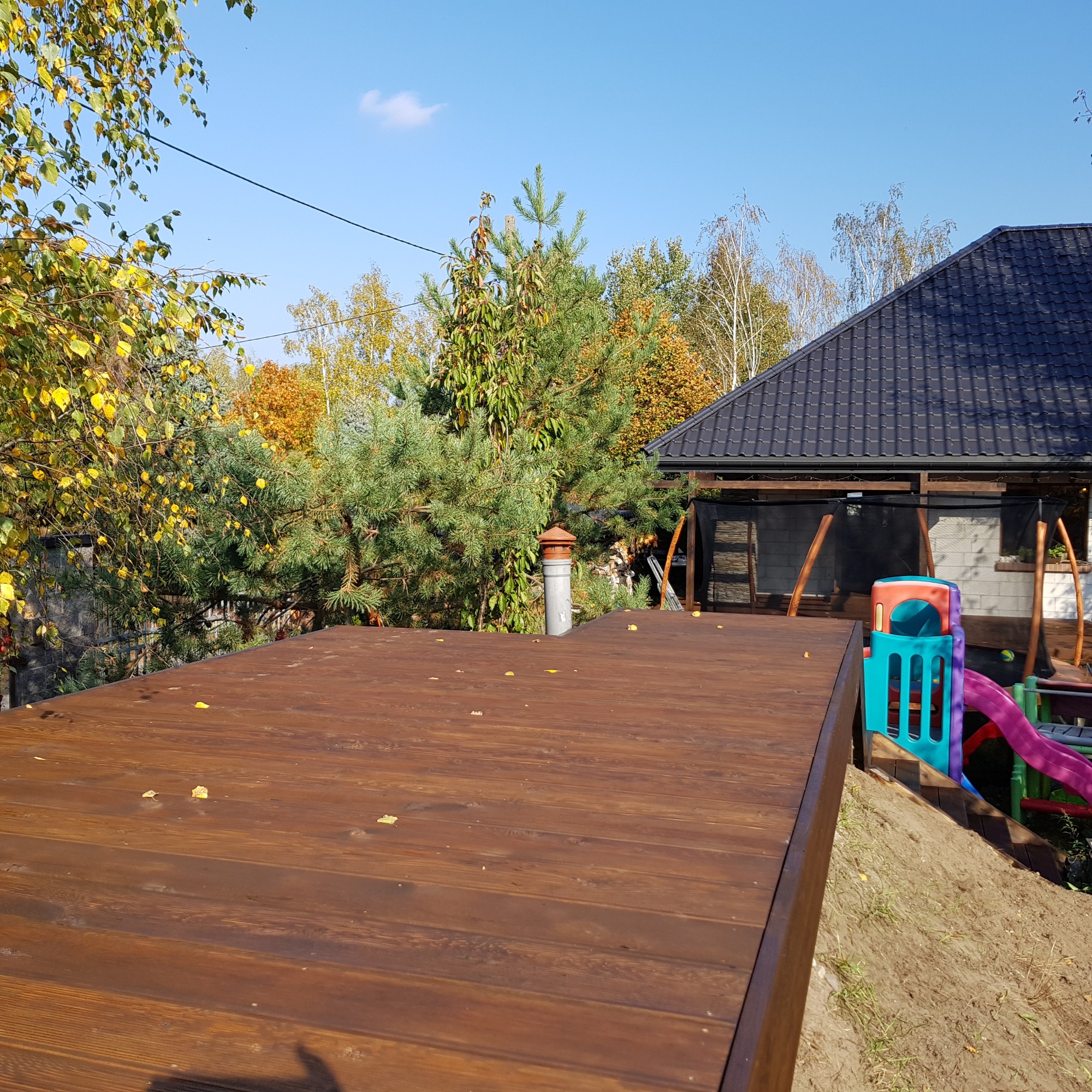 Brązowy taras z desek w Pruszkowie, z widocznym placem zabaw w tle i jesiennymi liśćmi na powierzchni.