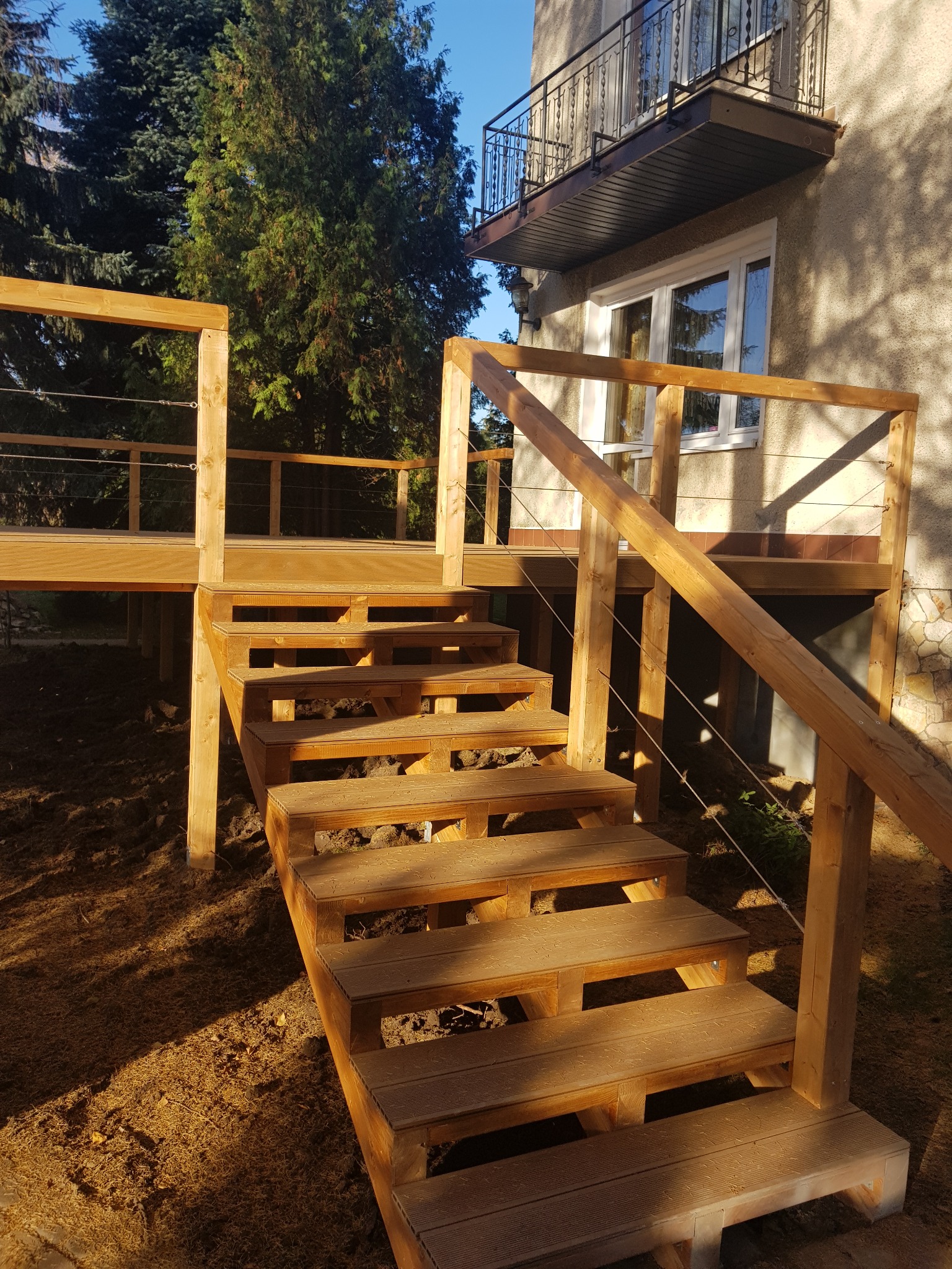 Drewniane schody prowadzące na podwyższony taras z balustradą z cienkich linek, widok z dołu na tle budynku z balkonem i drzew