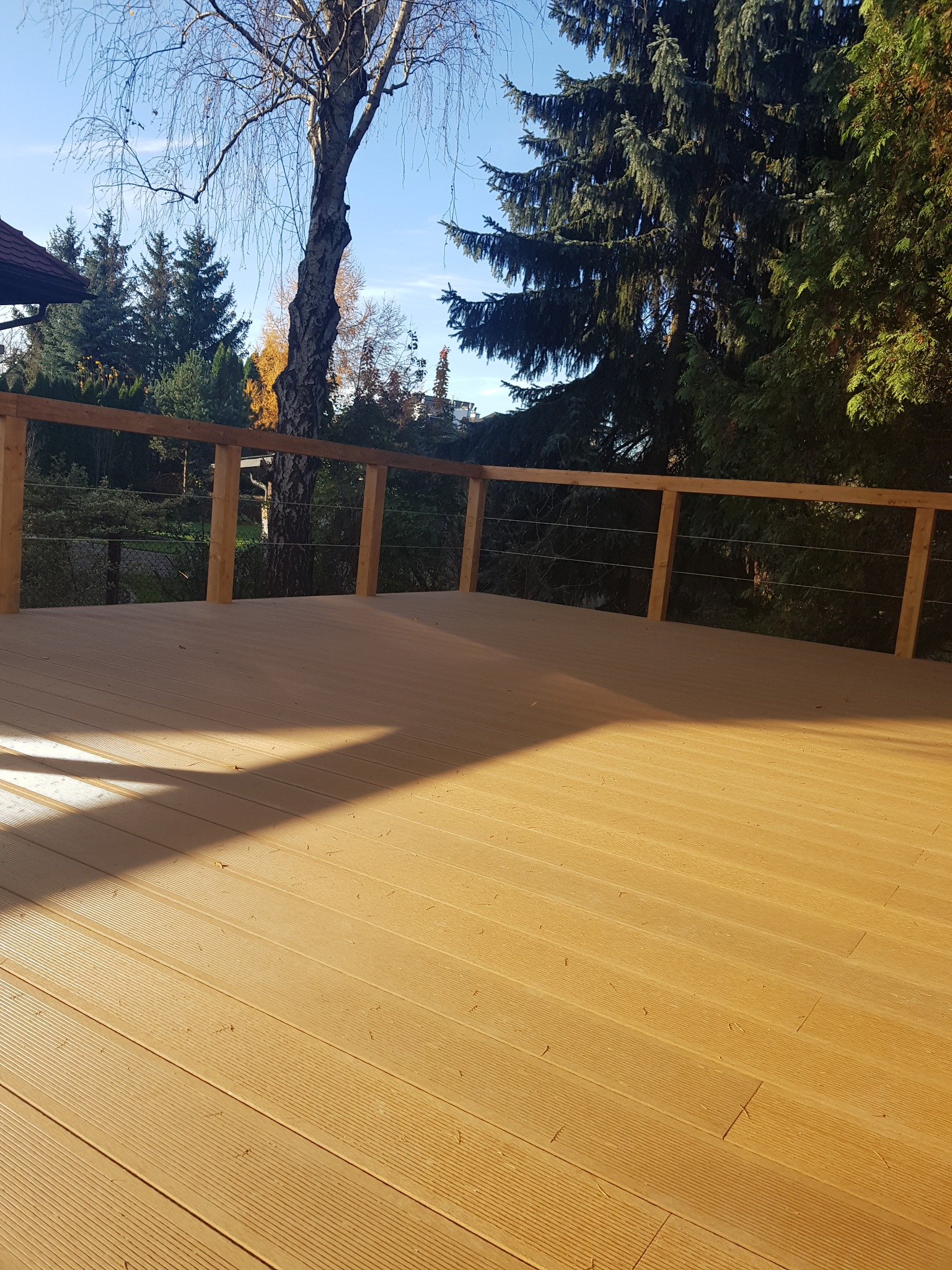 Jasna, nowa powierzchnia tarasu z desek kompozytowych z drewnianą balustradą i stalowymi linkami, w tle drzewa i błękitne niebo.