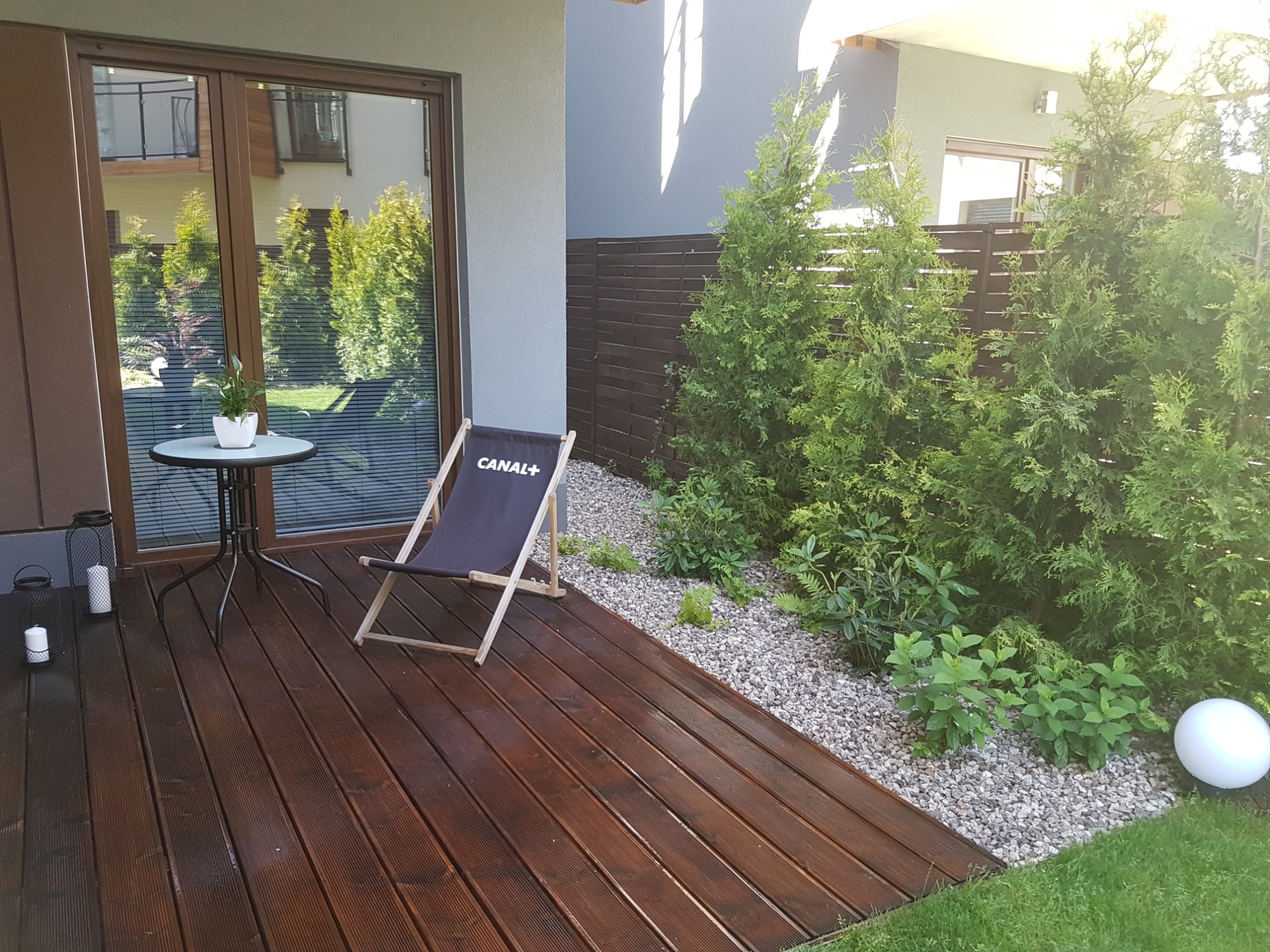 Ciemnobrązowy taras z desek, krzesło plażowe Canal+, stolik z kwiatem w doniczce, ozdobny lampion ze świecami, tuje i kamyczki, nowoczesny dom w tle.