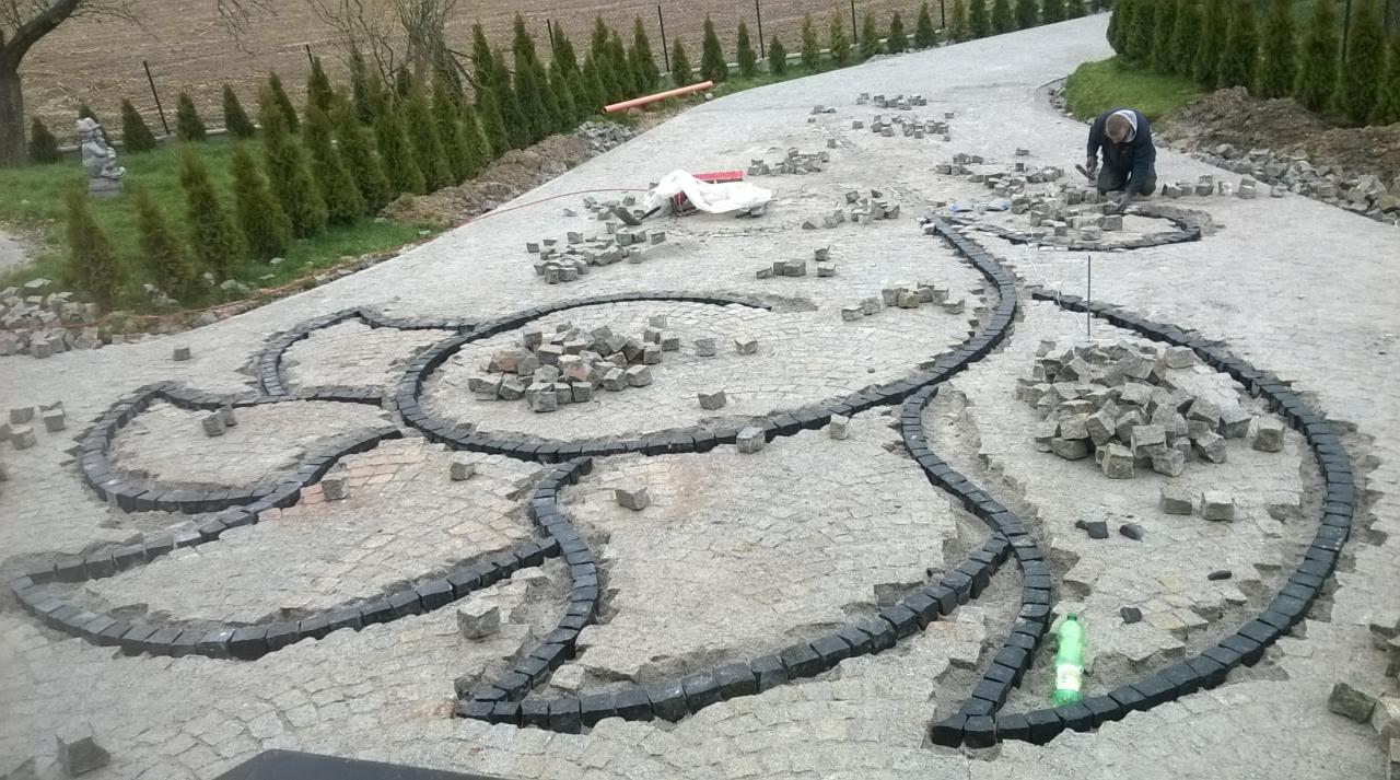 Układanie wzoru z kostki granitowej na podjeździe, widoczne obramowanie w kształcie płatków kwiatów i robotnik w trakcie pracy.