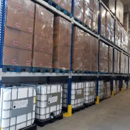 Wysokie regały magazynowe z paletami z kartonami owiniętymi folią i plastikowymi pojemnikami IBC z etykietami ostrzegawczymi na dole, stojące na betonowej posadzce.