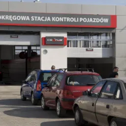 Budynek Okręgowej Stacji Kontroli Pojazdów w Braniewie z zaparkowanymi samochodami, widoczni mężczyzna z dzieckiem wchodzący do biura obsługi klienta.