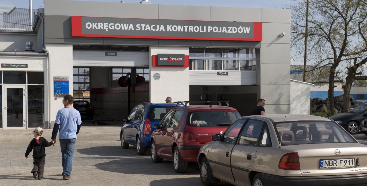 Budynek Okręgowej Stacji Kontroli Pojazdów w Braniewie z zaparkowanymi samochodami, widoczni mężczyzna z dzieckiem wchodzący do biura obsługi klienta.