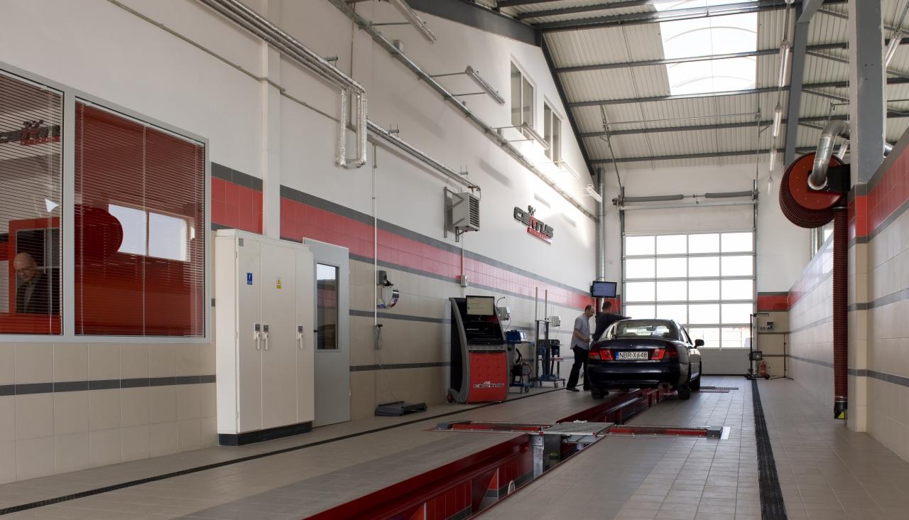 Samochód na stanowisku diagnostycznym w przestronnym warsztacie z dwoma pracownikami i urządzeniami pomiarowymi, widok z tyłu pojazdu.