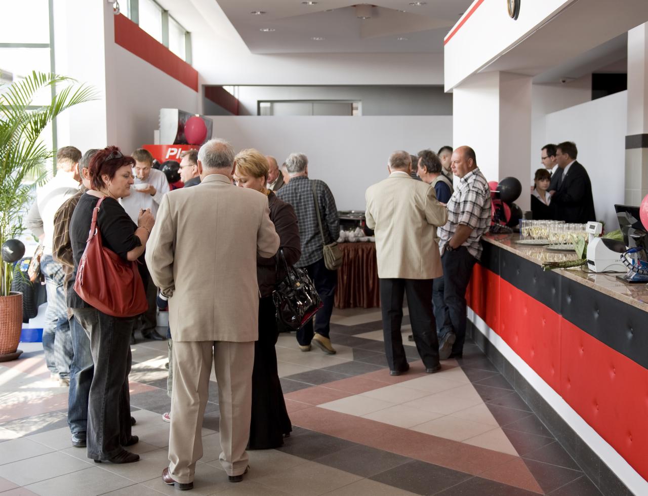 Wnętrze salonu samochodowego podczas uroczystości otwarcia lub jubileuszu, z grupą ludzi w różnym wieku, rozmawiających i stojących przy barze z szampanem.