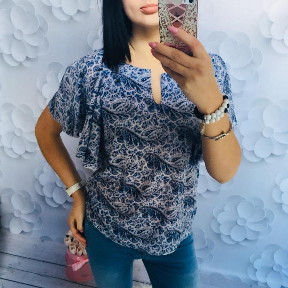 Kobieta robi selfie w bluzce z niebieskim wzorem paisley, jeansach i z bransoletkami na ręce, na tle ściany z papierowymi kwiatami.