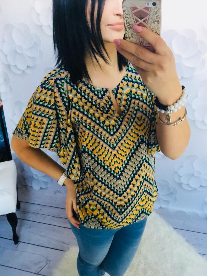 Kobieta robi selfie w bluzce z geometrycznym wzorem w odcieniach żółci, zieleni i granatu, z rozszerzanymi rękawami i małym wycięciem przy dekolcie, w tle dekoracja z białych, papierowych kwiatów.