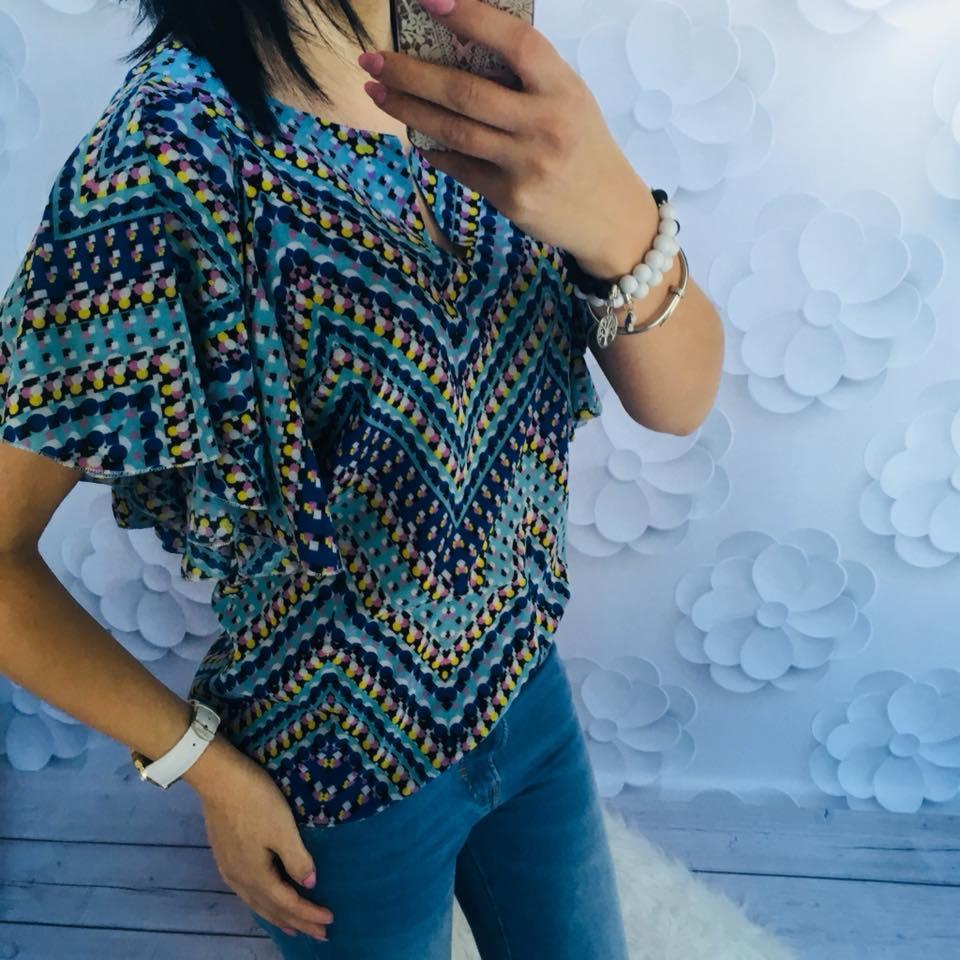 Kobieta robi selfie w bluzce z wzorzystym deseniem w odcieniach błękitu i granatu, jeansach i z bransoletkami na tle ściany z dekoracjami kwiatowymi.