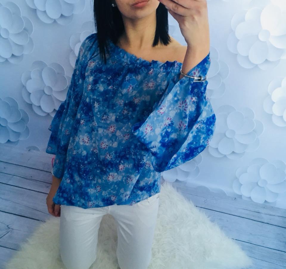 Kobieta pozuje w asymetrycznej, błękitnej bluzce w kwiatowy wzór z szerokimi rękawami i białych spodniach na tle dekoracji z papierowych kwiatów.
