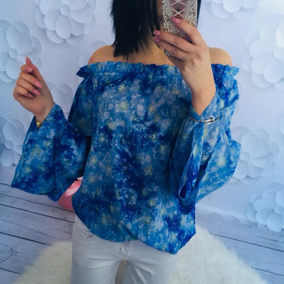 Kobieta robi selfie w bluzce z odkrytymi ramionami, w odcieniach błękitu z kwiatowym wzorem, na tle białych, papierowych kwiatów i drewnopodobnej podłogi.