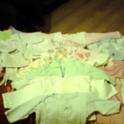 Różnorodny zestaw ubranek dla niemowląt w rozmiarze 56/62 cm, ułożonych na ciemnej powierzchni, widoczne body, pajacyki i kaftaniki w pastelowych kolorach, oświetlenie pomieszczenia tworzy cienie.