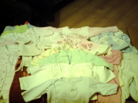 Różnorodny zestaw ubranek dla niemowląt w rozmiarze 56/62 cm, ułożonych na ciemnej powierzchni, widoczne body, pajacyki i kaftaniki w pastelowych kolorach, oświetlenie pomieszczenia tworzy cienie.