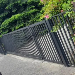 Czarna, przesuwna brama wjazdowa z pionowych profili na tle zieleni i fragmentu drewnianego ogrodzenia, widoczny fragment zaparkowanego samochodu.