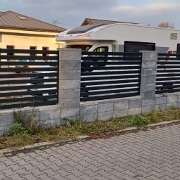 SB-Steel - Nowoczesne ogrodzenie panelowe z poziomych, czarnych profili i szarych, kamiennych słupków oraz podmurówki, montowane przez montera w czerwonej kurtce, na tle budynku i kampera.