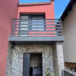 Balkon z poziomą szarą balustradą, elewacja budynku w odcieniach różu, kamienna okładzina wejścia, otwarte szare drzwi, dekoracje kwiatowe w donicach.