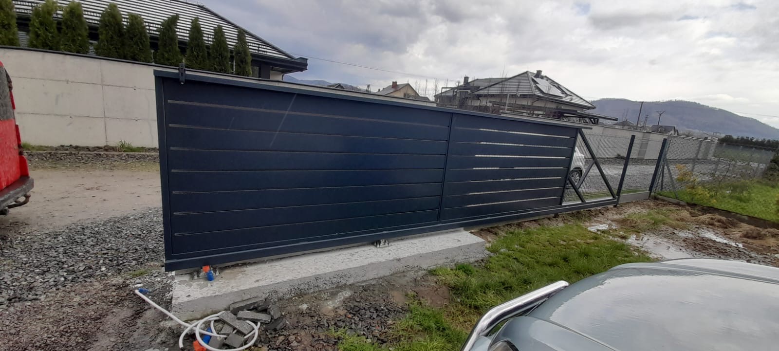 Nowoczesna, przesuwna brama wjazdowa z poziomych paneli, zamontowana na betonowej podstawie, widoczna z perspektywy ulicy, z domem i siatką ogrodzeniową w tle.