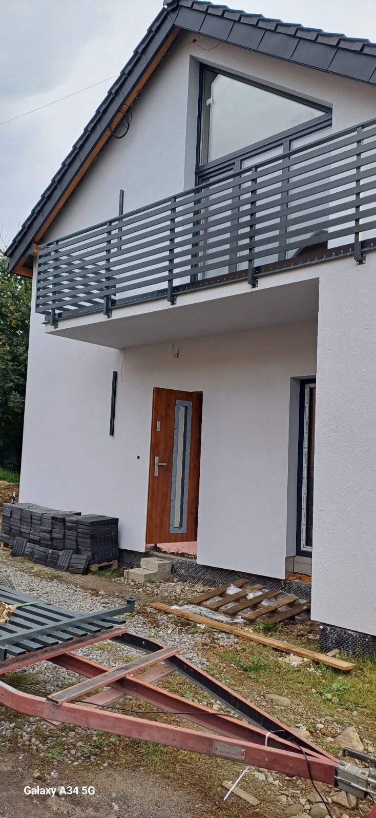 Nowoczesny dom z szarą, poziomą balustradą balkonową, widoczne materiały budowlane przed wejściem, w tym paleta z kostką brukową i rama przyczepki.