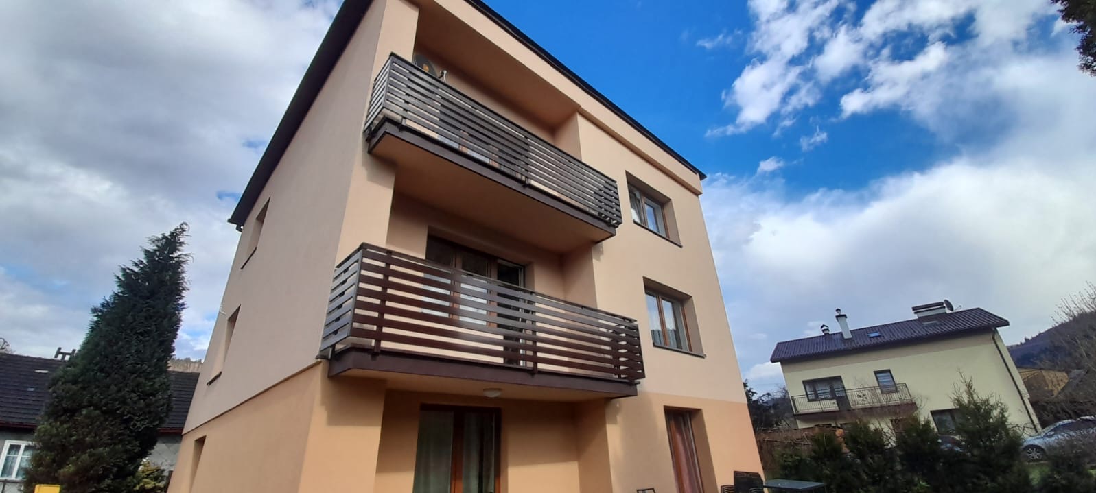 Nowoczesny, trzypiętrowy dom z beżową elewacją i balkonami z ciemnobrązowymi, poziomymi barierkami, na tle błękitnego nieba z chmurami.