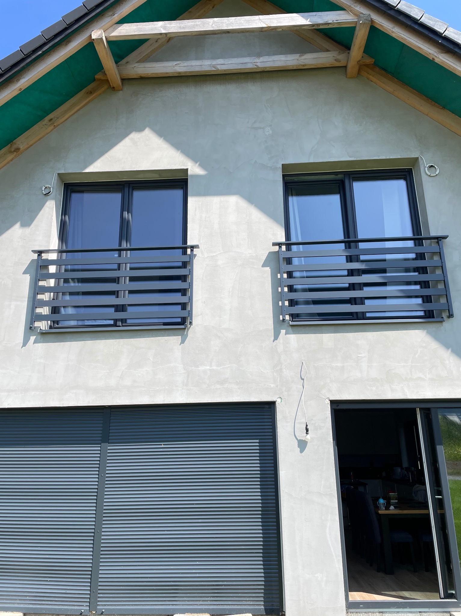 Elewacja domu z dwoma balkonami z poziomymi balustradami metalowymi i szarą roletą garażową.