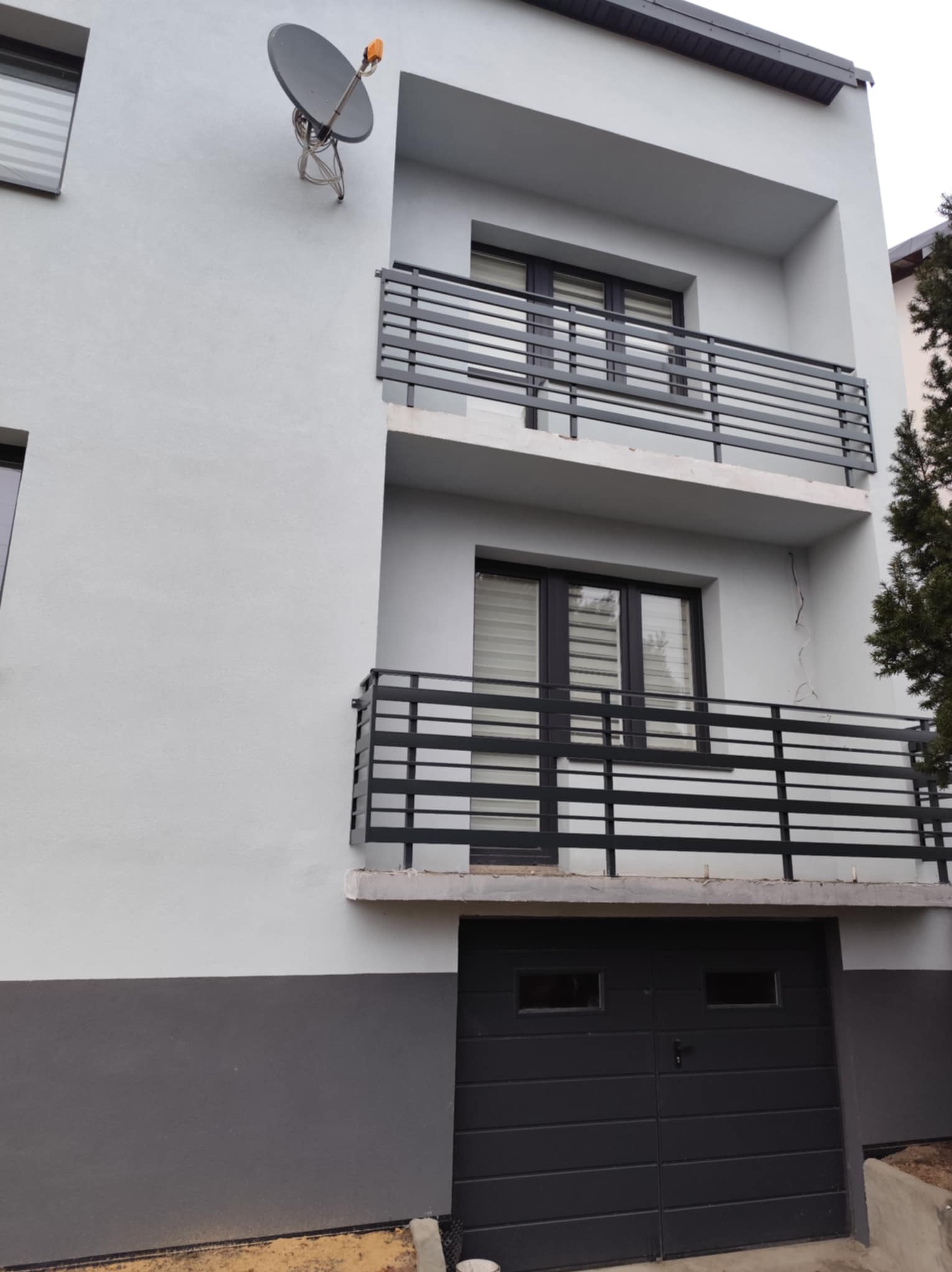 Dwupiętrowy dom z balkonami zabezpieczonymi poziomymi, czarnymi barierkami, antena satelitarna na elewacji i brama garażowa w kolorze grafitowym.