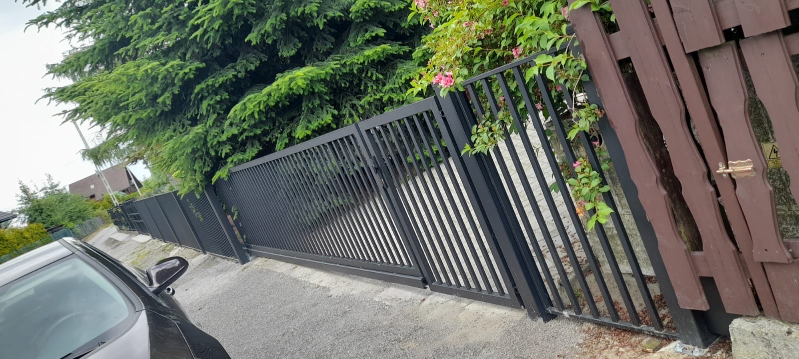 Czarna, przesuwna brama wjazdowa z pionowych profili na tle zieleni i fragmentu drewnianego ogrodzenia, widoczny fragment zaparkowanego samochodu.