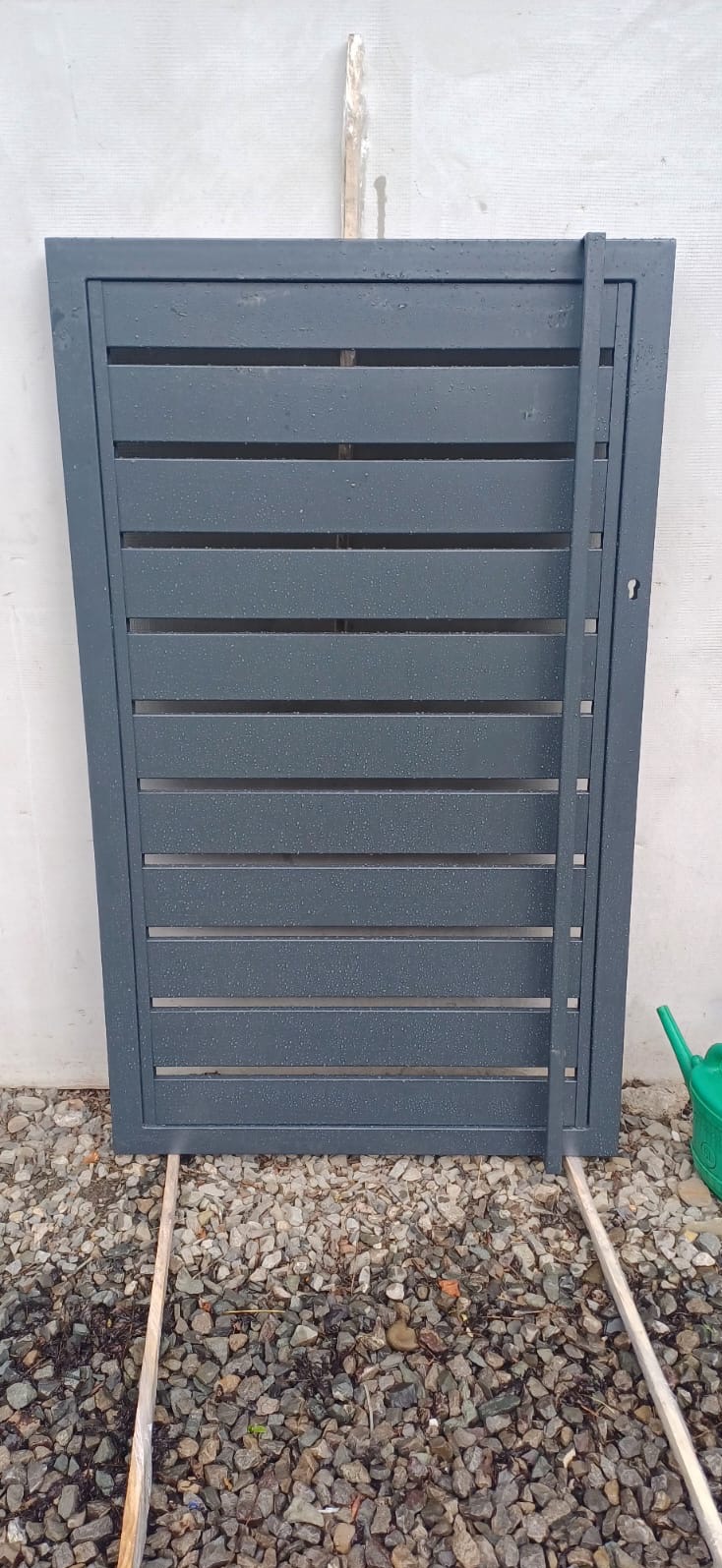 Szara metalowa furtka z poziomymi szczeblami, oparta o ścianę na żwirowym podłożu, widoczna zielona konewka.