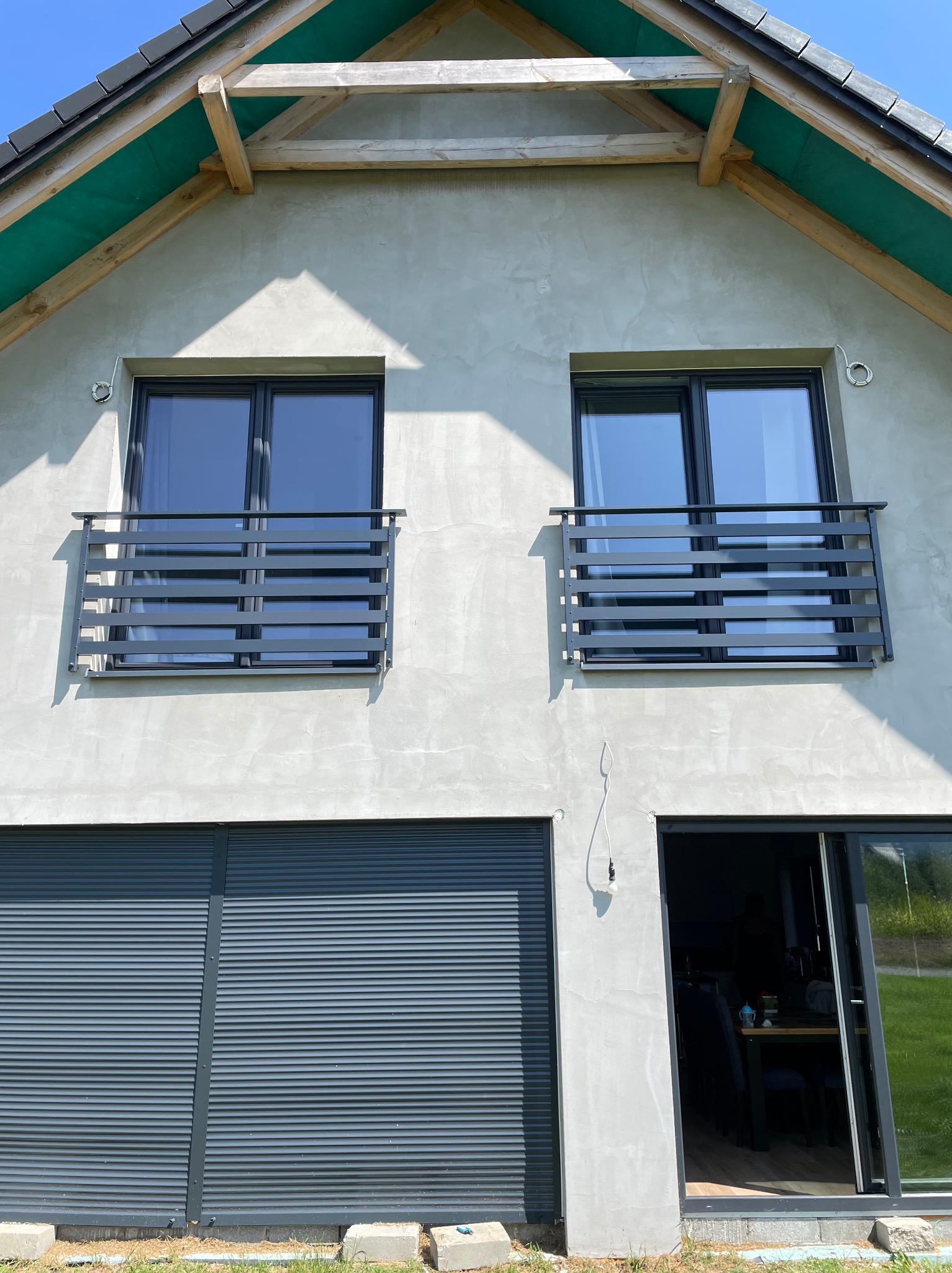 Nowoczesny dom z szarą elewacją, oknami z ciemnymi ramami i metalowymi barierkami balkonowymi oraz roletami zewnętrznymi w kolorze grafitowym.