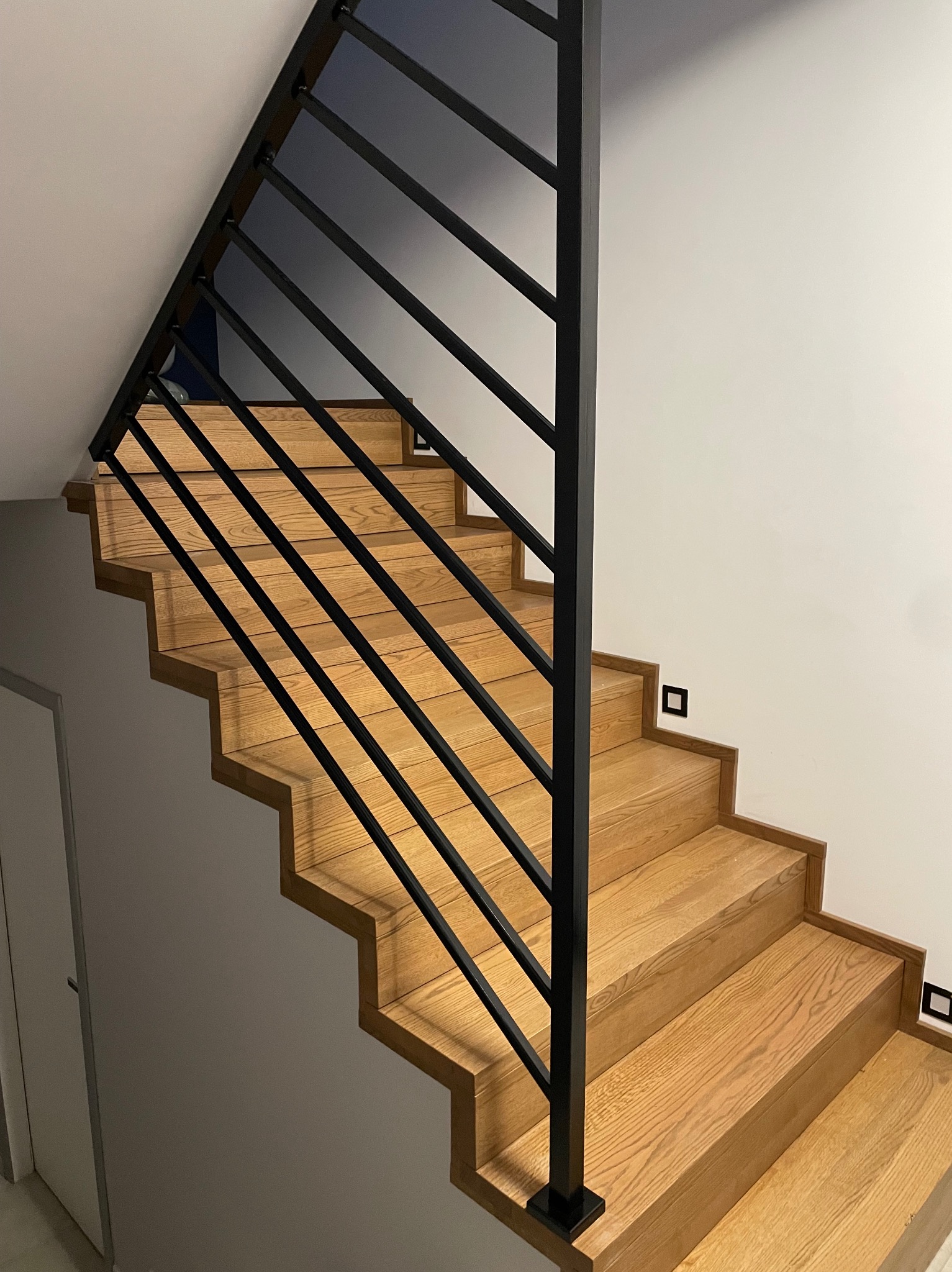 Schody drewniane z czarną, minimalistyczną barierką z metalowych prętów, widok z dołu, oświetlenie punktowe na ścianie.