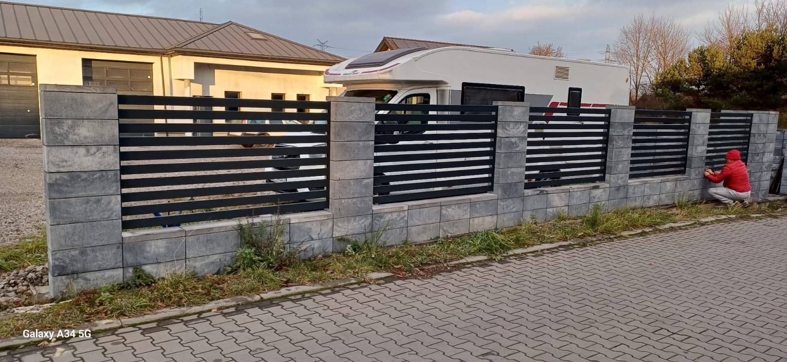 Nowoczesne ogrodzenie panelowe z poziomych, czarnych profili i szarych, kamiennych słupków oraz podmurówki, montowane przez montera w czerwonej kurtce, na tle budynku i kampera.