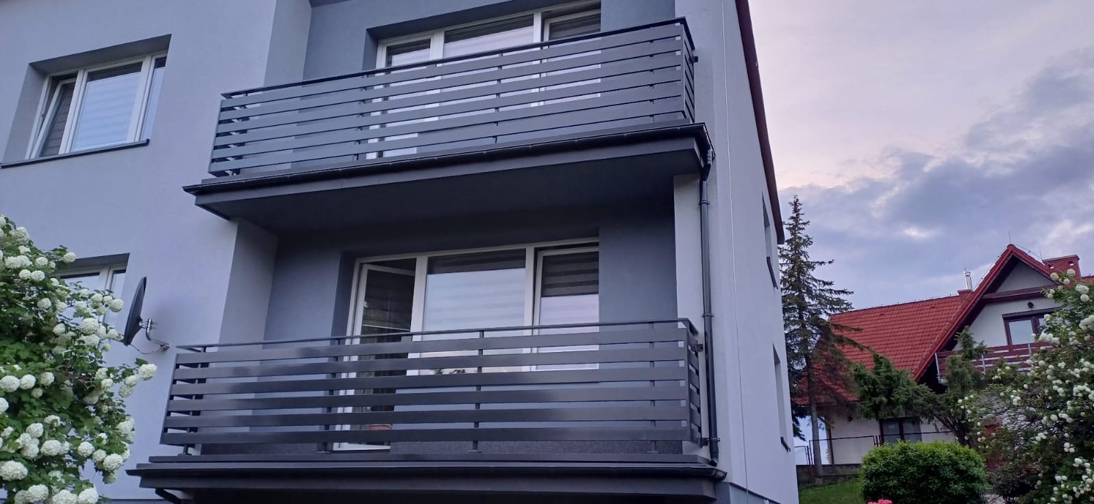 Dwupiętrowy budynek z szarymi balkonami z poziomymi, metalowymi barierkami, widok z dołu, z kwitnącymi krzewami w tle.