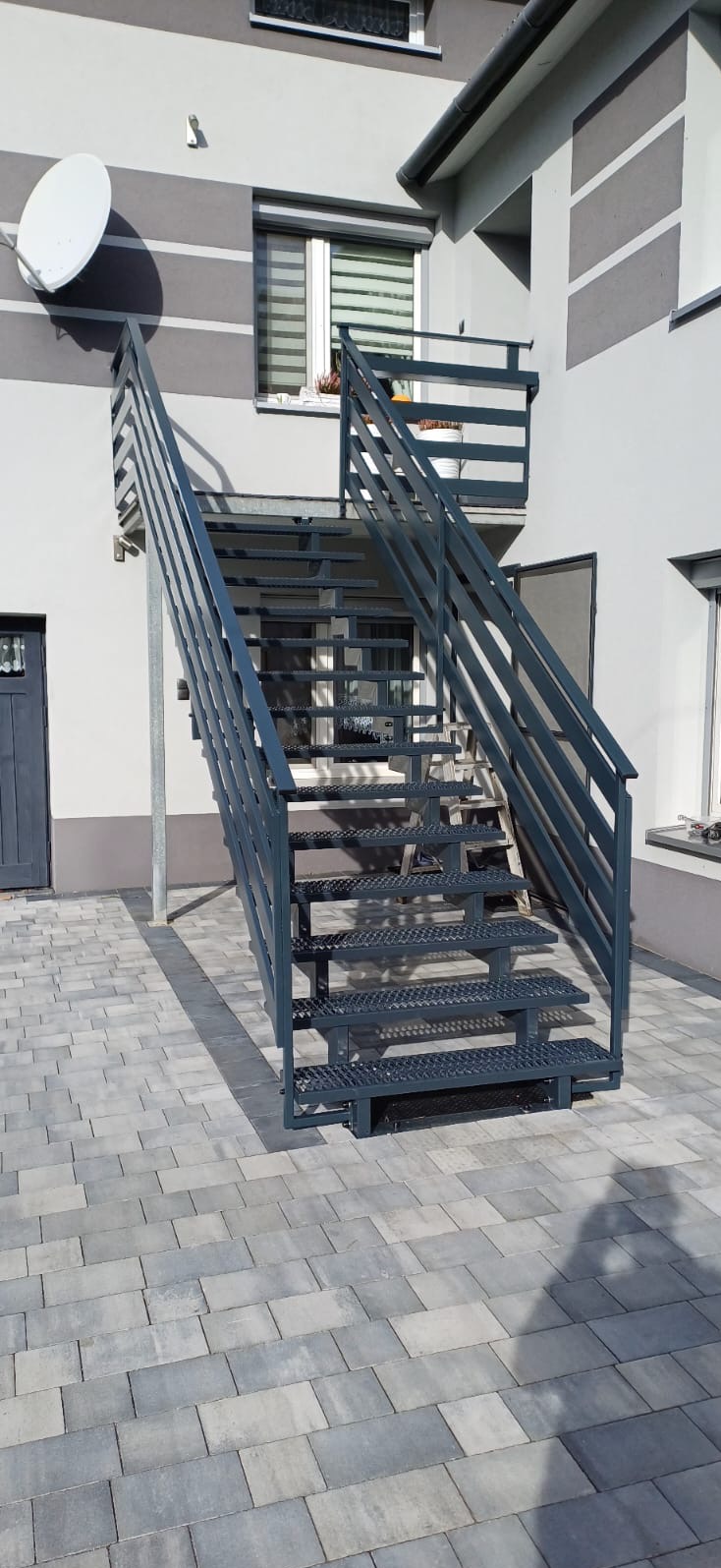 Zewnętrzne metalowe schody z ażurowymi stopniami i zabezpieczeniem bocznym, prowadzące do balkonu w nowoczesnym domu z szarą elewacją.