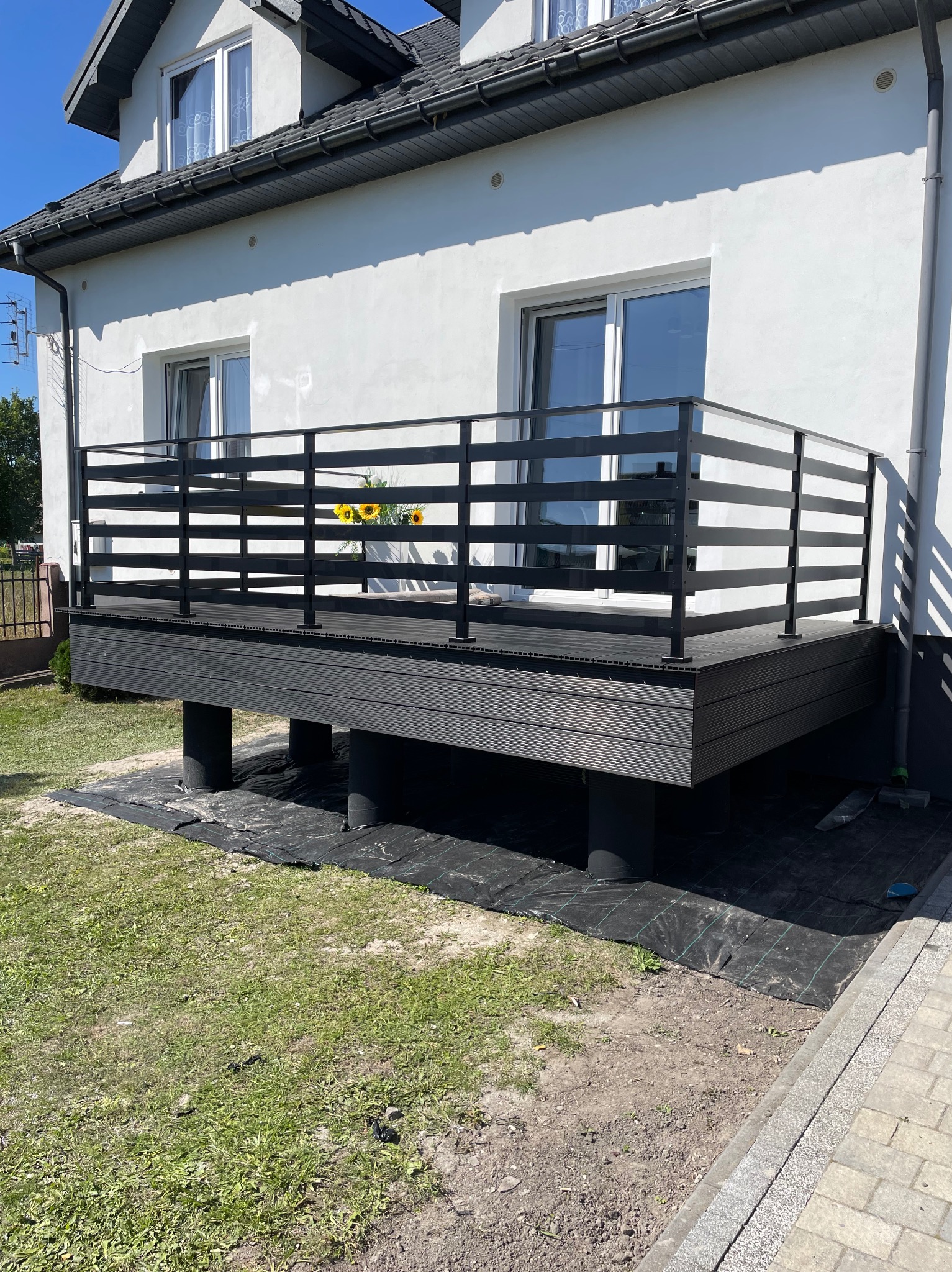 Nowoczesny balkon z czarną, poziomą balustradą i kompozytową podłogą, widok z zewnątrz. Kwiaty w wazonie na balkonie, biała elewacja budynku w tle.