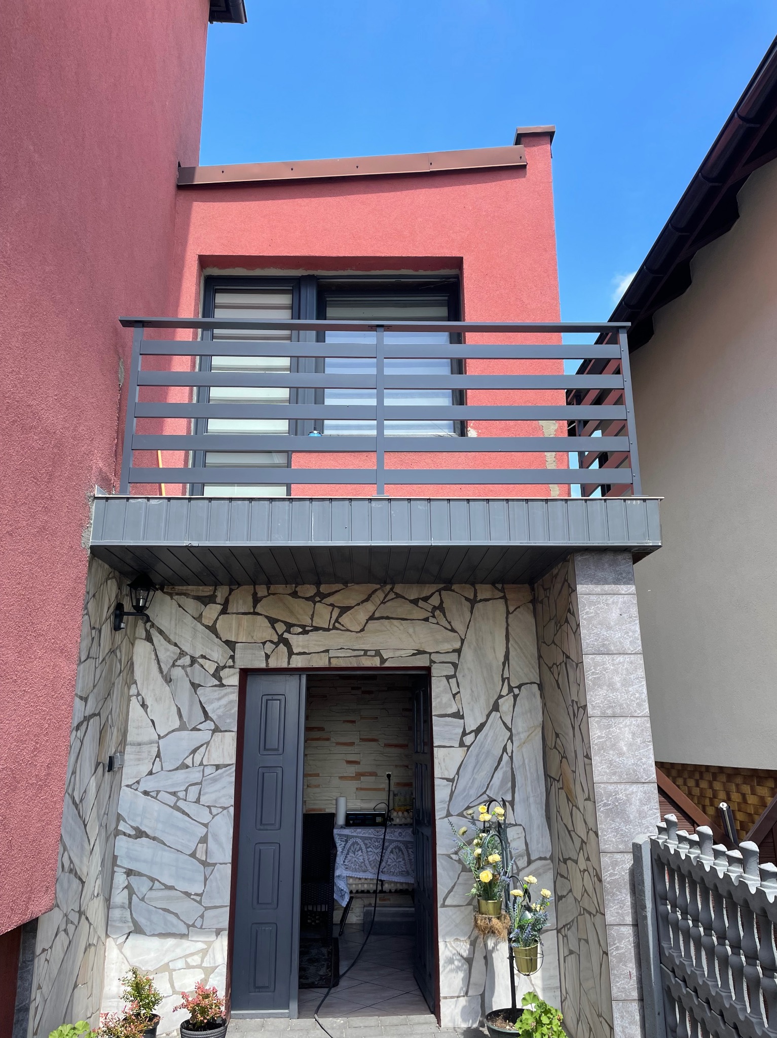 Balkon z poziomą szarą balustradą, elewacja budynku w odcieniach różu, kamienna okładzina wejścia, otwarte szare drzwi, dekoracje kwiatowe w donicach.