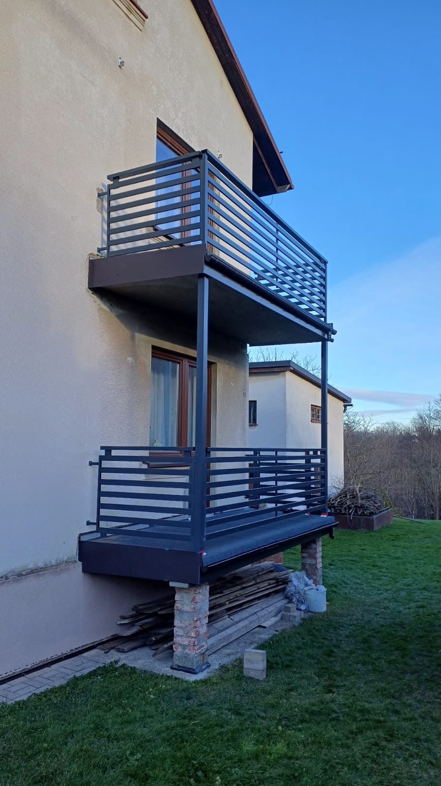 Dwupoziomowy balkon z prostymi, metalowymi barierkami w kolorze grafitowym, mocowany do elewacji domu, widoczne ceglane podpory i składowane deski u podstawy.