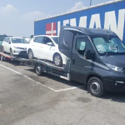 Szara laweta Iveco z dwoma białymi samochodami osobowymi na przyczepie, zaparkowana na parkingu z niebieską naczepą w tle.